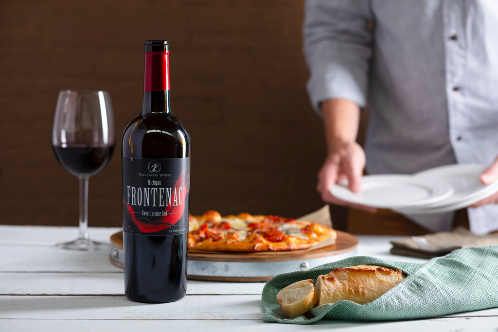 A man sets the table with plates, a bottle of Tanglewood Winery's Frontenac Red Wine, pizza, and bread.